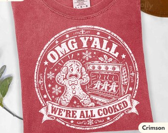 May include: A red short-sleeve t-shirt featuring a white circular graphic. The design shows a gingerbread person with a shocked expression standing next to an open oven containing three smaller gingerbread cookies. Text reads OMG Y'ALL WE'RE ALL COOKED.
