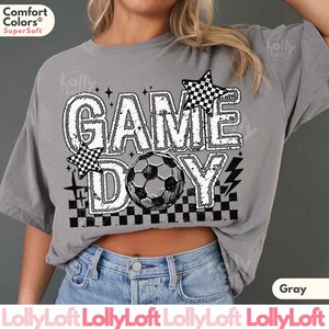 May include: Gray t-shirt with the words "GAME DAY" in a white, outlined font, featuring a soccer ball and checkered star accents. The shirt is made of a soft material and is a comfortable fit.