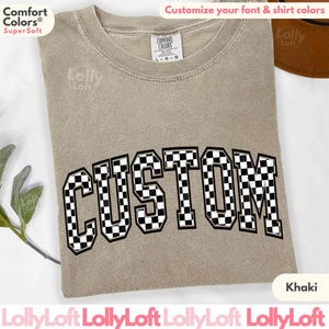 May include: Khaki t-shirt with the word "CUSTOM" in black and white checkered font. The shirt is made of a soft, comfortable fabric.
