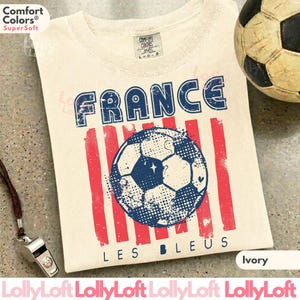 May include: Ivory-colored t-shirt with a distressed graphic featuring the word "FRANCE" in blue, red stripes, and a soccer ball design. The text "LES BLEUS" is below the soccer ball. A soccer ball and a whistle are in the background.