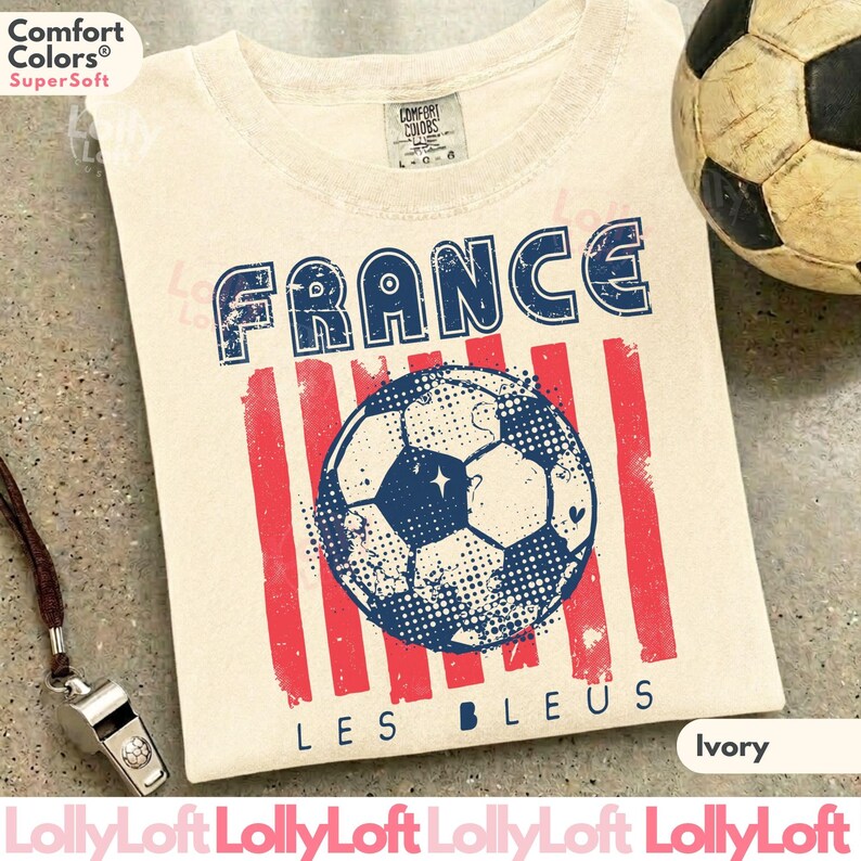 May include: Ivory-colored t-shirt with a distressed graphic featuring the word "FRANCE" in blue, red stripes, and a soccer ball design. The text "LES BLEUS" is below the soccer ball. A soccer ball and a whistle are in the background.