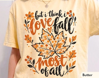 May include: A pale yellow t-shirt featuring a graphic print with the text "but i think i love fall most of all" in black and orange script. The design includes a large central maple leaf with a polka dot pattern and orange floral accents.
