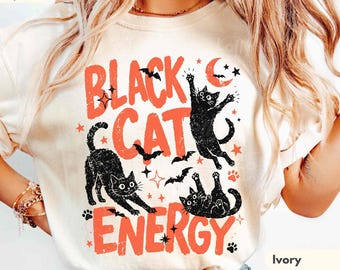 May include: An ivory t-shirt featuring the orange text "BLACK CAT ENERGY" and illustrations of three black cats in various poses, surrounded by small black bats, orange stars, and a crescent moon. The shirt has a crew neck and short sleeves.
