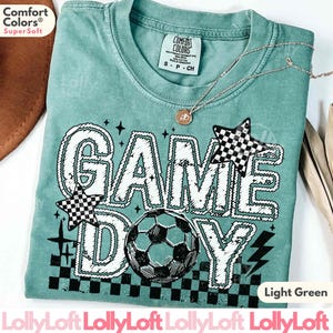 May include: Light green Comfort Colors t-shirt with "GAME DAY" graphic, soccer ball, and checkered star accents. Features a crew neck and short sleeves. Includes a gold necklace. The shirt is labeled "Super Soft".