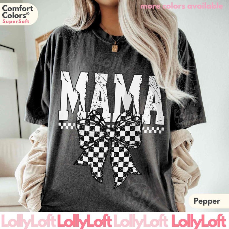 Op de afbeelding: Donkergrijs t-shirt met het woord "MAMA" in versleten witte letters. Een zwart-wit geblokte strik staat eronder. Het shirt heeft het Comfort Colors Super Soft logo.