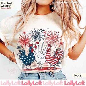 Pode incluir: Uma camiseta branca com um gr&aacute;fico de tr&ecirc;s galos com designs de bandeira americana e fogos de artif&iacute;cio. O texto "Comfort Colors&reg; Super Soft" est&aacute; na parte superior da camiseta. A camiseta est&aacute; dispon&iacute;vel em marfim.