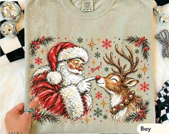 May include: A light beige t-shirt featuring a graphic of Santa Claus touching the nose of a reindeer. The text Boop is printed in red. The design includes green pine branches, red berries, and red and gold snowflakes on a Comfort Colors label.