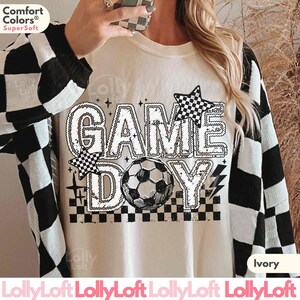 May include: Ivory-colored sweatshirt with the words "GAME DAY" in a bold, outlined font, featuring a soccer ball and checkered star accents. The sweatshirt has a crew neck and long sleeves. The sleeves are black and white checkered.