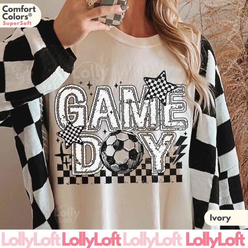 May include: Ivory-colored sweatshirt with the words "GAME DAY" in a bold, outlined font, featuring a soccer ball and checkered star accents. The sweatshirt has a crew neck and long sleeves. The sleeves are black and white checkered.