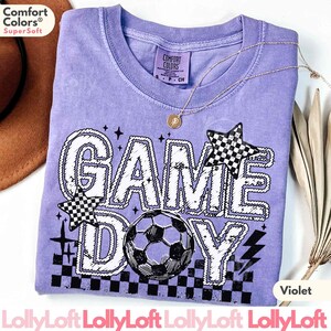 May include: Violet-colored t-shirt with the words "GAME DAY" in a white, outlined font. The design includes a soccer ball, checkered stars, and lightning bolts. The shirt has a crew neck and a small tag that says "Comfort Colors".