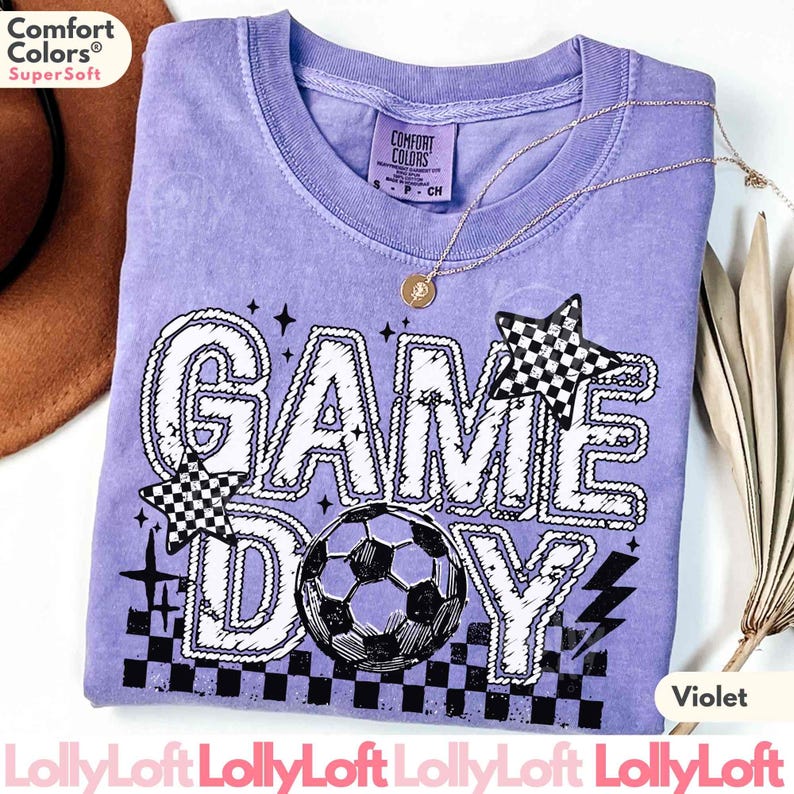 May include: Violet-colored t-shirt with the words "GAME DAY" in a white, outlined font. The design includes a soccer ball, checkered stars, and lightning bolts. The shirt has a crew neck and a small tag that says "Comfort Colors".