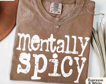 May include: Espresso-colored Comfort Colors t-shirt with the phrase "mentally spicy" in white, distressed lettering. The shirt has a crew neck and short sleeves. A silver necklace with a small pendant rests on the shirt. The shirt is labeled "Espresso & White."