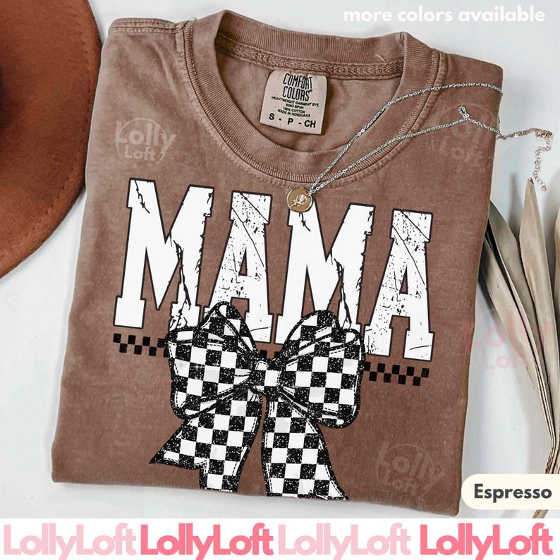 Op de afbeelding: Espresso-kleurig t-shirt met het woord "MAMA" in grote witte, versleten letters. Een zwart-wit geblokte strik is onder het woord gecentreerd. Het shirt heeft een Comfort Colors-label en een zilveren ketting.