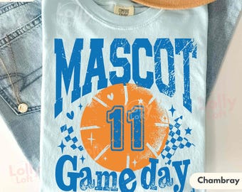 May include: Light blue t-shirt with a distressed blue graphic that reads "MASCOT Game day 11" over an orange basketball. The shirt is made of Comfort Colors Super Soft material.