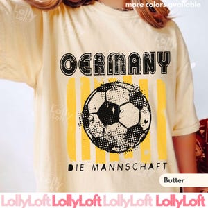 May include: Butter-colored t-shirt with a distressed graphic featuring the word "GERMANY" above a soccer ball and vertical yellow stripes. The text "DIE MANNSCHAFT" is below the ball. The shirt is a casual style.