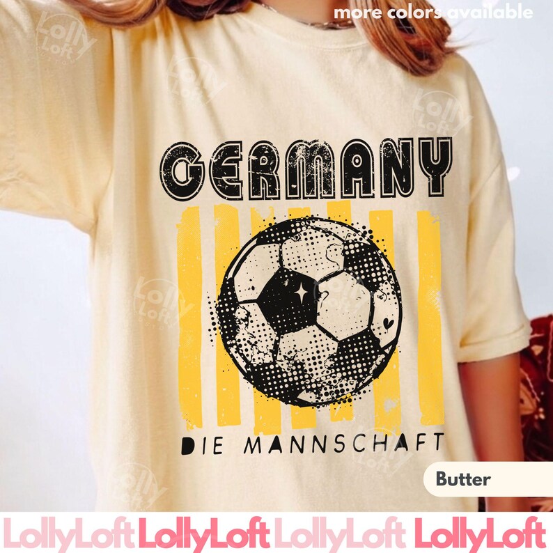 May include: Butter-colored t-shirt with a distressed graphic featuring the word "GERMANY" above a soccer ball and vertical yellow stripes. The text "DIE MANNSCHAFT" is below the ball. The shirt is a casual style.
