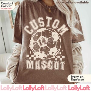 Op de afbeelding: Een bruin t-shirt met korte mouwen en een grote crèmekleurige afbeelding van een voetbal met het nummer 14 in het midden. De tekst CUSTOM MASCOT is in een vet, verweerd lettertype boven en onder de voetbalafbeelding gedrukt.