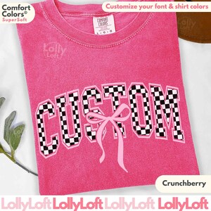 May include: A pink Comfort Colors Super Soft t-shirt with the word "CUSTOM" in a black and white checkered pattern. A pink bow is below the word. The shirt also has the text "Crunchberry" and "Customize your font & shirt colors".