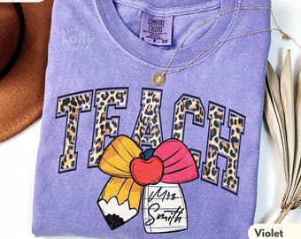 May include: Violet Comfort Colors t-shirt with the word "TEACH" in leopard print letters. A pencil, apple, and bow graphic is below the word. The shirt also has the text "Mrs. Smith" on a notepad.