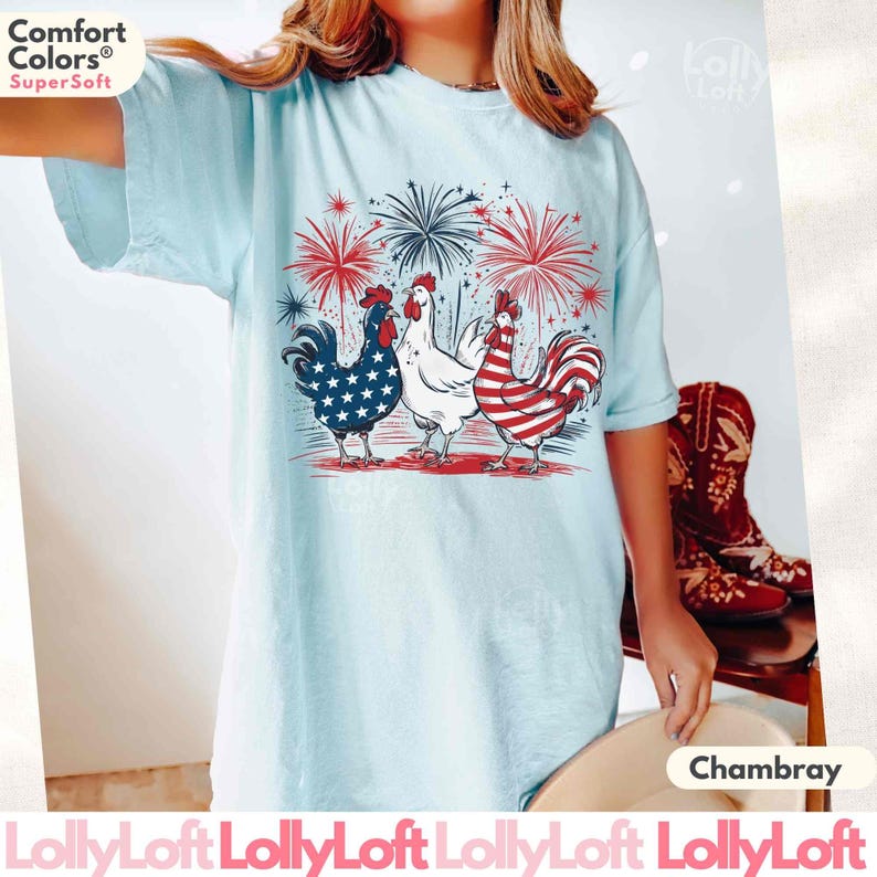 Pode incluir: Uma camiseta azul claro com um gr&aacute;fico de tr&ecirc;s galos usando as cores da bandeira americana. Os galos est&atilde;o cercados por fogos de artif&iacute;cio. O texto "Comfort Colors SuperSoft" est&aacute; no canto superior esquerdo da imagem. O texto "Chambray" est&aacute; no canto inferior direito da imagem.