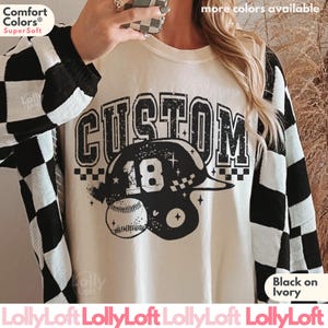 May include: An ivory t-shirt featuring a black graphic print with the word CUSTOM in a distressed font above a baseball helmet displaying the number 18 and a baseball. The sleeves have a black and white checkered pattern, and the shirt is ivory.