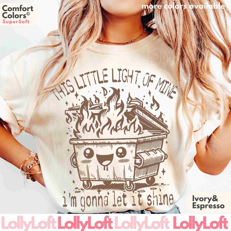 Pu&ograve; includere: Una maglietta beige con una grafica marrone di un cassonetto della spazzatura in fiamme con un viso sorridente e il testo "This little light of mine, I'm gonna let it shine".
