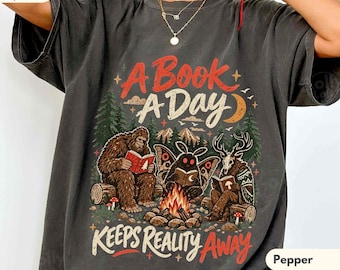 May include: A charcoal grey t-shirt featuring a graphic print of a Bigfoot, a mothman, and a deer-headed creature reading books around a campfire in a forest. The text reads A BOOK A DAY KEEPS REALITY AWAY. The shirt has short sleeves and a crew neckline.