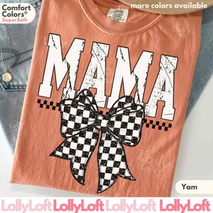 Op de afbeelding: Een yam-kleurig t-shirt met het woord "MAMA" in versleten witte letters. Een grote zwart-wit geblokte strik is onder het woord gecentreerd. De shirt heeft een ronde hals en korte mouwen. De merknaam "Comfort Colors" is zichtbaar.