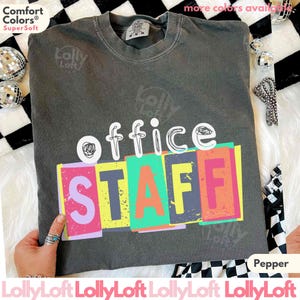 May include: A dark gray t-shirt with the words "office STAFF" in a colorful, retro design. The shirt is made of a soft material and has a crew neck. The text "Comfort Colors Super Soft" is in the upper left corner.
