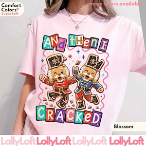May include: A light pink t-shirt featuring a graphic of two nutcracker figures with cracked heads. The text reads "AND then I CRACKED" in colorful block letters. The design includes small star accents and a pink wavy border around the nutcrackers.