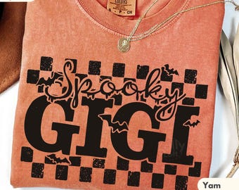 May include: A burnt orange short-sleeved t-shirt featuring a black checkered pattern background with the text Spooky GIGI in a bold, stylised font. The design includes small black bat silhouettes scattered throughout the graphic.