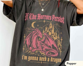 May include: A charcoal grey short-sleeve t-shirt featuring a graphic print of a pink dragon in front of a castle. The text reads If The Horrors Persist I'm gonna need a dragon. The shirt has a crew neckline and a relaxed fit.