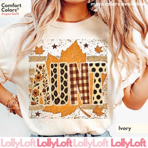 May include: An ivory t-shirt featuring a large graphic of a maple leaf with the word AUNT printed across it. The letters are filled with floral, leopard print, plaid, and polka dot patterns. The design includes small stars and distressed textures.