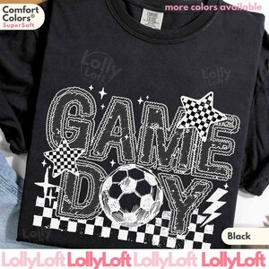 May include: Black t-shirt with a white graphic that says "GAME DAY" with a soccer ball in the center. The design includes stars and a checkered pattern. The shirt is made of a soft material.