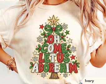 May include: An ivory t-shirt featuring a graphic of a Christmas tree formed by the text "OH COME LET US ADORE HIM" in red, green, and white. The tree is decorated with a gold star, green bows, red berries, and white snowflakes.