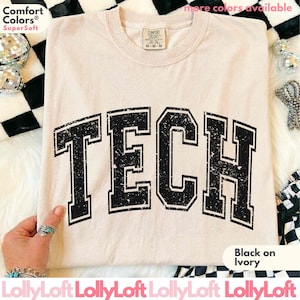 May include: A short-sleeved ivory t-shirt featuring the word TECH printed in a large, distressed black collegiate font across the chest. The shirt has a crew neckline and a Comfort Colors brand label on the inside of the collar.