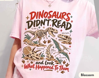May include: A light pink t-shirt featuring a graphic print of dinosaur skeletons including a T-rex, Triceratops, and Pterodactyl. The text reads Dinosaurs Didn't Read and Look What Happened To Them, with labels for T-rex, Velociraptor, Neck Vertebrae, and Pterodactyl.