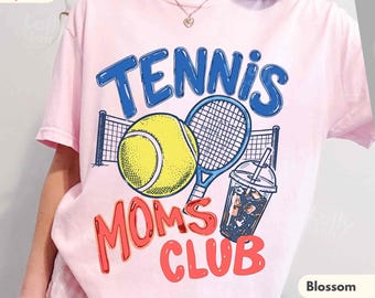 May include: A light pink t-shirt featuring a graphic print with the text TENNIS MOMS CLUB in blue and red. The design includes a yellow tennis ball, a blue tennis racket, a net, and a clear plastic cup with a straw and dark liquid.