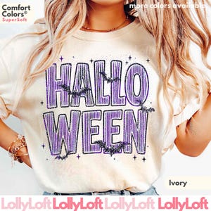 May include: An ivory t-shirt featuring the purple text HALLOWEEN in a striped font. The design includes small black bat silhouettes and star accents scattered around the letters. The shirt has a Comfort Colors SuperSoft label at the top left.