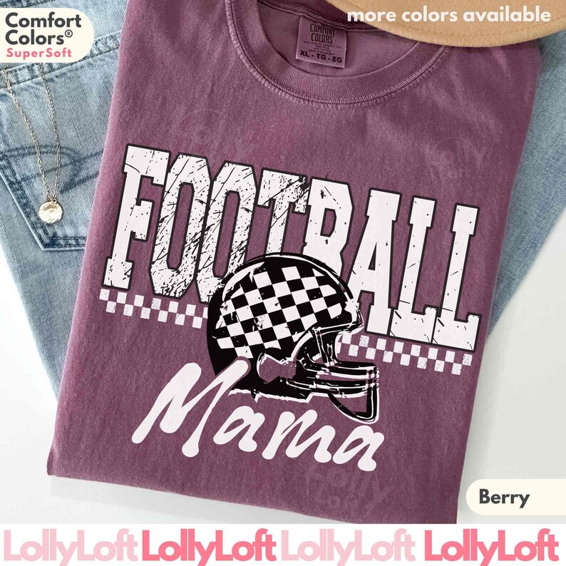 May include: Berry-colored t-shirt with the words "FOOTBALL Mama" in white, featuring a football helmet with a checkered design. The shirt is a Comfort Colors Super Soft tee.