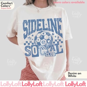 May include: Cream-colored t-shirt with a blue graphic that reads "SIDELINE SOCIAL CLUB" with a soccer ball illustration. The shirt is made of a soft material and has a relaxed fit. The text "Denim on White" is also visible.