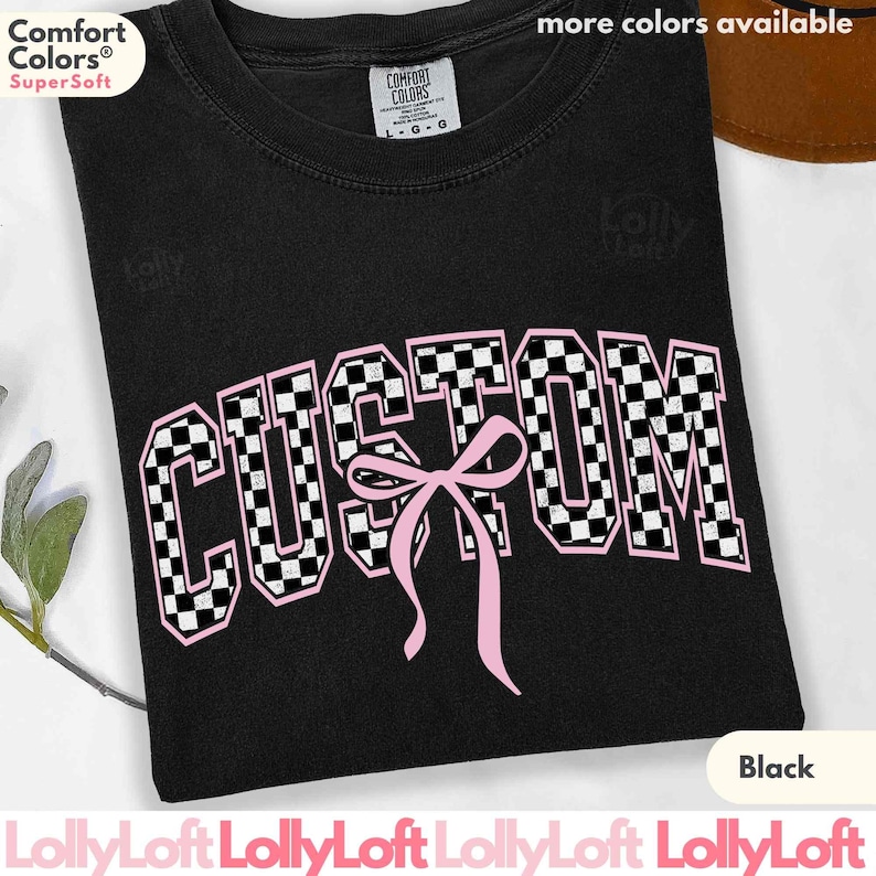 May include: Black t-shirt with the word "CUSTOM" in a pink and black checkered design, with a pink bow. The shirt has the Comfort Colors Super Soft logo. The text "more colors available" is at the top.