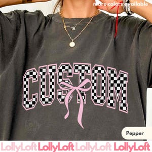 May include: Dark gray sweatshirt with the word "CUSTOM" in a checkered pattern with a pink outline and a pink bow. The text "more colors available" is at the top. The word "Pepper" is at the bottom right.