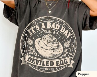 May include: A charcoal grey t-shirt featuring a circular graphic with a central illustration of a deviled egg. The text reads IT'S A BAD DAY TO BE A DEVILED EGG, surrounded by decorative leaf and star motifs in a distressed, off-white ink.