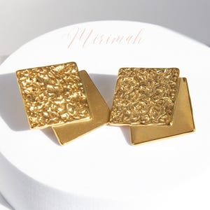 Puede incluir: Un par de pendientes de color dorado con un diseño geométrico texturizado. Cada pendiente tiene dos cuadrados superpuestos.