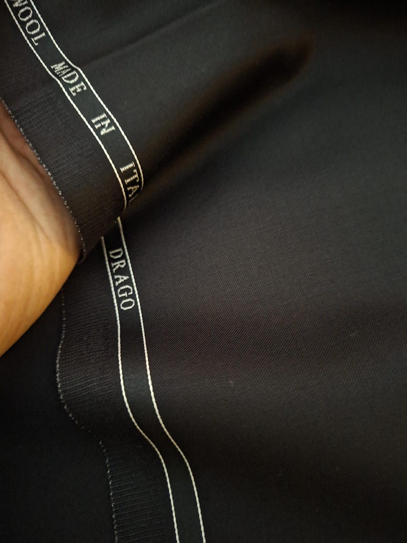 Puede incluir: Primer plano de tela de lana negra con una franja blanca que dice "MADE IN ITALY" y "DRAGO". La tela est&aacute; doblada, mostrando su textura y brillo. El material parece de alta calidad, adecuado para la sastrer&iacute;a.