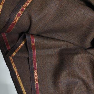 May include: Close-up of brown fabric with a herringbone pattern. The fabric has a red and gold trim with the text "Dormeuil" and "Made in England" on it.