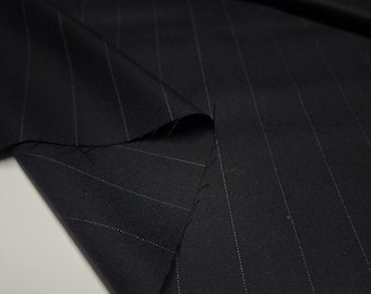 May include: A piece of dark navy blue fabric featuring a subtle pinstripe pattern with thin, light-colored vertical lines. The textile is folded, creating soft drapes and shadows that highlight the smooth texture and the consistent, repeating stripe design.