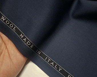 May include: A swatch of navy blue wool fabric. The fabric is smooth and appears to be high-quality. A black label with white text reads "WOOL MADE IN ITALY BY TRA". The fabric is held by a hand.