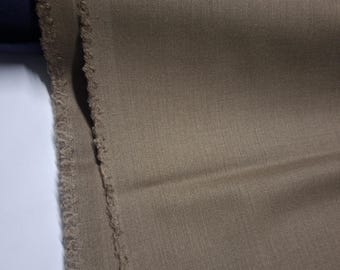 May include: Close-up of a tan fabric with a textured surface. The fabric has a raw, frayed edge on the left side. The material is folded, showing the weave and color variations. A roll of dark blue fabric is visible in the background.